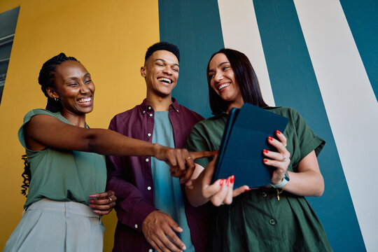 Diverse young colleagues laughing while looking at tablet during casual meeting in office