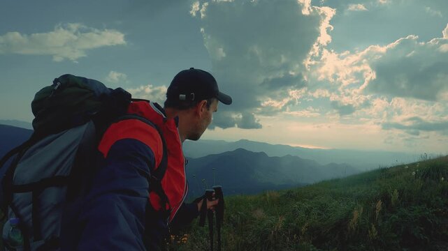 A man wearing a baseball cap, red and blue windbreaker and a backpack walks along a grassy hillside holding trekking poles and looking at distant rolling mountains under a dramatic sky
