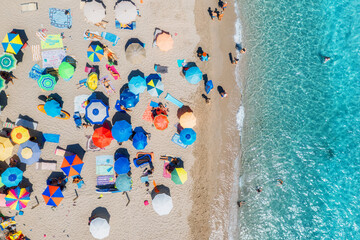 Aerial view of colorful umbrellas on sandy beach, swimming people in blue sea on sunny summer day....