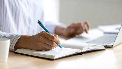 Close-up of a person's hand writing in a notebook near a laptop, showcasing a calm and focused work-from-home atmosphere, ideal for productivity, planning, and journaling concepts.