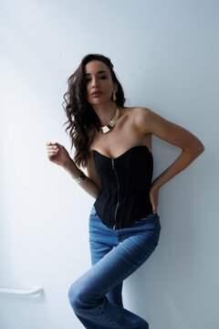 Beautiful tall thin brunette with curly hair in living room. The girl in black korset and jeans, room background. Studio photo shoot for a model. Portrait.