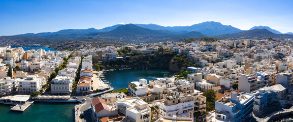 Aerial view of Agios Nikolaos, Crete, featuring Voulismeni Lake, whitewashed buildings, narrow streets, a canal, and rugged mountains under a clear sky. © Aerial Film Studio