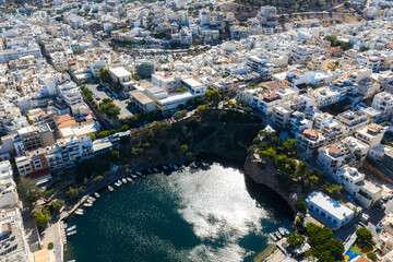 Aerial view of Agios Nikolaos, Crete, featuring Voulismeni Lake, whitewashed buildings, narrow streets, and surrounding hilly terrain under sunlight. © Aerial Film Studio