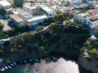 Aerial view of Agios Nikolaos, Crete, showing a rocky coastline, a small harbor with docked boats, whitewashed houses, and lush greenery. © Aerial Film Studio