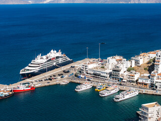 Aerial view of Agios Nikolaos, Crete, showing a cruise ship docked at the harbor, smaller boats, whitewashed buildings, and distant hills across the sea. © Aerial Film Studio