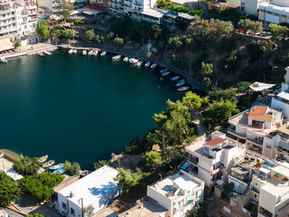 Aerial view of Agios Nikolaos, Crete, featuring a circular lake, whitewashed buildings, terracotta roofs, small boats, and natural cliffs. © Aerial Film Studio