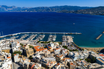 Fototapeta premium Aerial view of Agios Nikolaos, Crete, showing a marina with boats, white and pastel buildings, red tiled roofs, blue sea, and distant mountains.