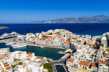 Naklejka premium Aerial view of Agios Nikolaos, Crete, showing a harbor with colorful buildings, small boats, a marina with yachts, and mountains in the background.