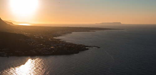 Aerial view of Stalida town on Crete's coastline during sunset. Calm sea waters shimmer under golden light, with a distant island on the horizon.