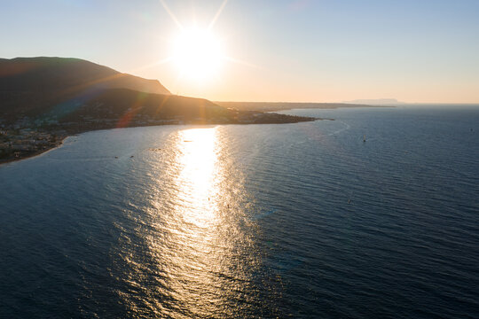 Aerial view of Crete Island's coastline during sunset, with golden reflections on the sea, mountains on the left, and a small town by the shore. - Powered by Adobe