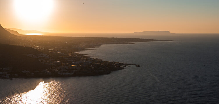 Aerial view of Stalida town on Crete's coastline during sunset. Calm sea waters shimmer under golden light, with a distant island on the horizon.