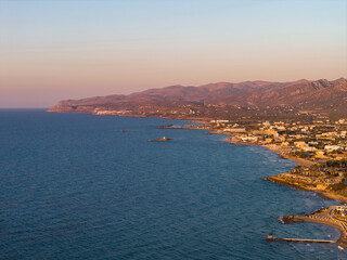 Fototapeta premium Aerial view of Stalida, Crete, showing the Aegean Sea, rugged coastline, small beaches, and town buildings with hills and mountains at sunset.