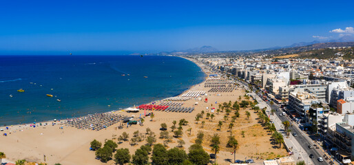 Aerial view of Rethymno town in Crete, Greece, featuring a sandy beach with sun loungers, blue Mediterranean waters, urban buildings, and distant mountains. © Aerial Film Studio