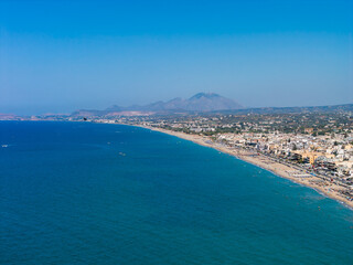 Fototapeta premium Aerial view of a coastal town on Crete Island, Greece, featuring sandy beaches, vibrant blue waters, rugged mountains, and a clear blue sky.
