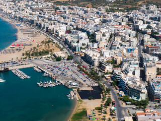 Fototapeta premium Aerial view of Rethymno, Crete, showing a marina with boats, a sandy beach with sunbeds, a promenade with buildings, and green hills in the background.