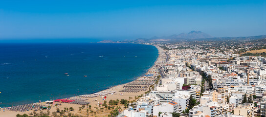 Aerial view of Rethymno town in Crete, Greece, showing a sandy beach with sunbeds, turquoise sea, whitewashed buildings, and hills in the background. © Aerial Film Studio