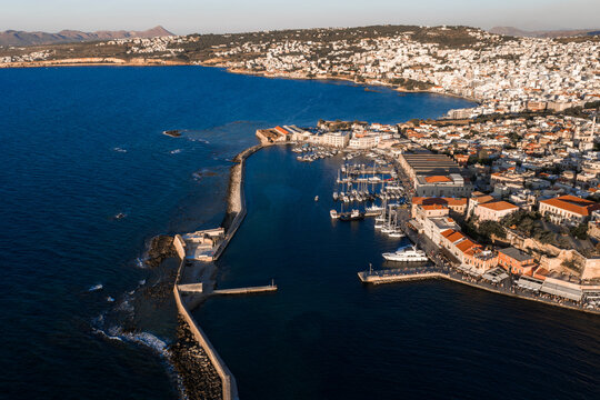 Aerial view of Chania, Crete, showcasing the Venetian harbor, docked boats, red tiled roofs, light colored buildings, and distant mountains. - Powered by Adobe