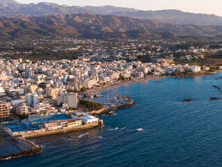Aerial view of Chania, Crete, Greece, at sunset. White and pastel buildings line the shoreline, with piers, boats, turquoise waters, and mountains visible.