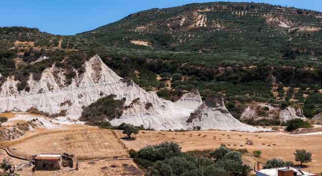 Komolithi white hills in Potamida, Crete, Greece, with conical formations, olive groves, terraced hills, a dry field, hay bales, and a rustic building.