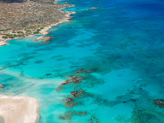 Obraz premium Aerial view of Elafonisi Beach in Crete, Greece, showing turquoise waters, darker coral patches, sandy shoreline, and sparse vegetation.