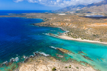 Obraz premium Aerial view of Elafonisi Beach in Crete, Greece, showing turquoise and blue waters, shallow lagoons, rocky outcrops, and distant mountains under clouds.