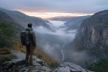 Fototapeta premium Man hiker overlooking foggy canyon valley with winding river at sunrise or sunset. Adventure and travel concept.