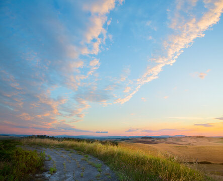 Beautiful Italian summer mountain countryside landscape with dirt road and cloudy sunset sky along the way; Sunrise panorama over summer golden field.