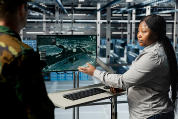 Teamworking army experts in data center confirming target locations using aerial image overlay on monitor. Military units in server room gathering strategic terrain data via satellite systems