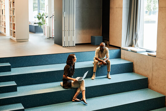 Horizontal high angle view long shot of young African American man and Caucasian woman sitting on stair bench in modern library using Internet, drinking coffee and chatting