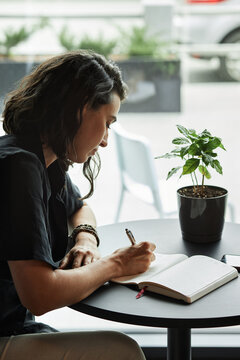 Vertical medium side view portrait shot of beautiful young Caucasian woman sitting at cafe table writing something in her notebook