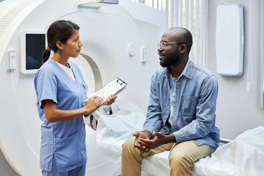 Horizontal shot of Hispanic medical specialist working in modern hospital standing in front of African American patient listening to him and making notes in papers