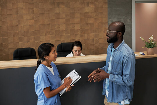 Horizontal medium long shot of African American man standing near hospital reception desk asking question to doctor