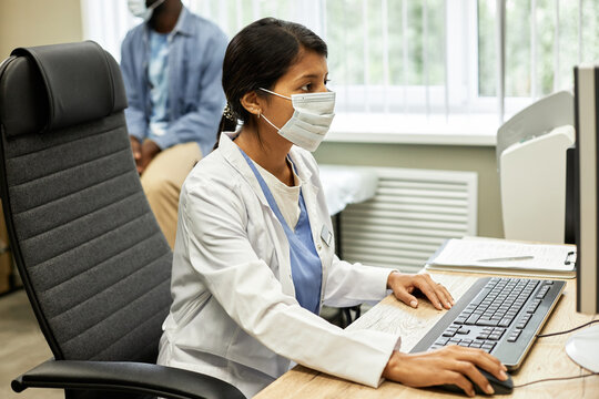 Horizontal medium shot of unrecognizable woman wearing mask and white coat working on computer in modern hospital