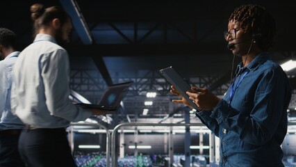 Server room admin using tablet and headset microphone to provide infrastructure tech support. Data center IT specialist resolves backend issues while coordinating with remote teams,