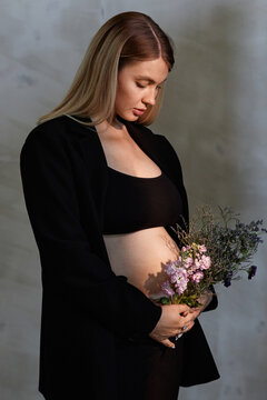 Vertical medium studio portrait of lovely young Caucasian woman expecting child wearing stylish black outfit holding flowers looking at tummy