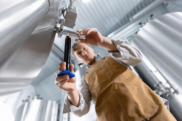 Caucasian male adult brewer testing beer sample in brewery with stainless steel tanks, Quality control concept