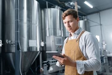 Male brewer using tablet in modern brewery with large steel tanks