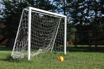 White soccer goal with net and colorful ball on grass field in park setting with trees background, outdoor sports recreation area. Concept of sports equipment, youth programs, recreational facilities.