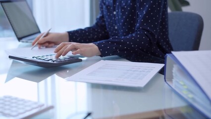 A female accountant with blue dotted blousy is using a laptop computer and calculator to calculate...