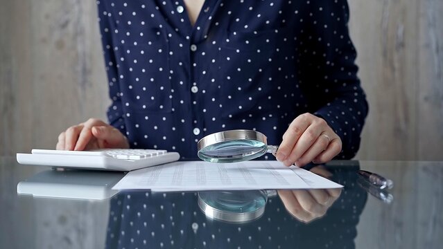 Auditor woman analyzing financial documents with magnifying glass and calculator at her office desk in formal dark blue blouse with polka dots. Business people concept - Powered by Adobe