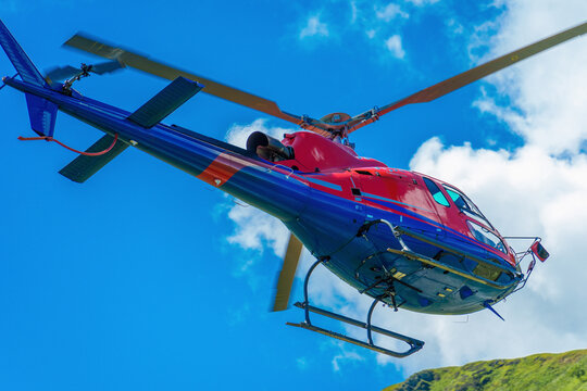 bright red helicopter flying against a clear blue sky, showcasing the majestic Swiss Alps in the background with fluffy white clouds