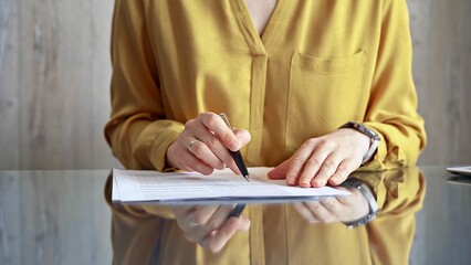 Businesswoman in yellow blouse is signing corporate contract at her office desk in formal white blouse with polka dots. Business people concept