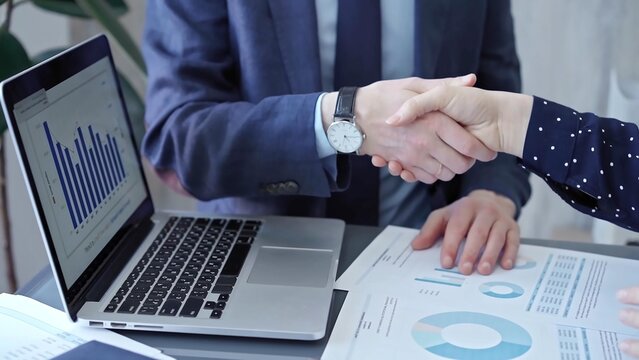 Two business people professionals shaking hands in office setting, sealing a deal with laptop and financial graphs visible. Business handshake over laptop and reports - Powered by Adobe