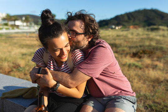 A man embraces his partner with love, while they spend time at the sun, sitting on a walk, in a coastal area, while drinking beer - Powered by Adobe