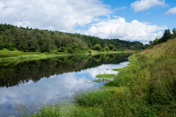 Russian countryside river flowing gently between green banks under cloudy summer sky. Natural landscape with forest reflections, calm water, and peaceful rural scenery.