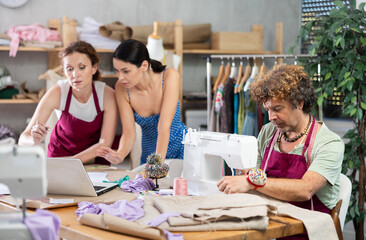 Two female dressmakers discussing dress designs on laptop. Man works on a sewing machine