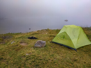 Obraz premium Green tent on misty lake shore with a fireplace nearby. Foggy camping scene, mysterious atmosphere, outdoor adventure in nature.