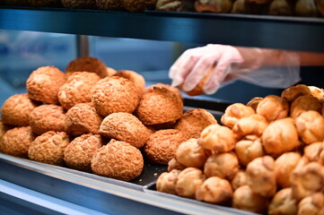 Professional bakery worker wearing gloves while handling fresh cream puffs. Popular French-origin pastry with crispy shell and creamy filling displayed in commercial bakery setting.
