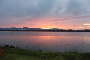 Fototapeta premium A breathtaking sunset over a calm river with vibrant orange and purple hues painting the sky. A small boat glides across the water, leaving gentle ripples under the silhouette of distant mountains.
