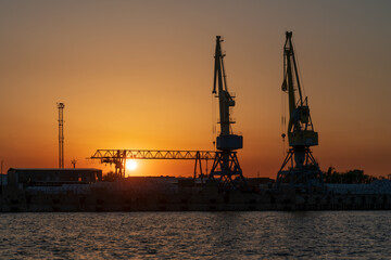 Obraz premium Port cranes for cargo transshipment on the mooring line of the Astrakhan port on the right bank of the Volga River against the background of the setting sun, Astrakhan, Russia